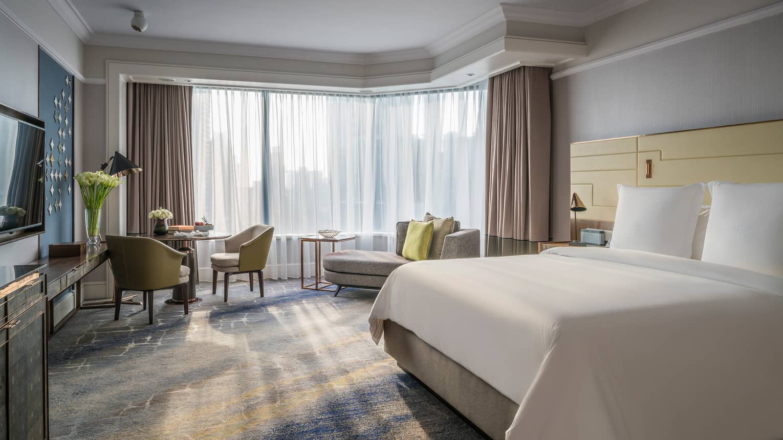 Sunny Deluxe Room bed with white linens, small dining table and chaise lounge, white curtains