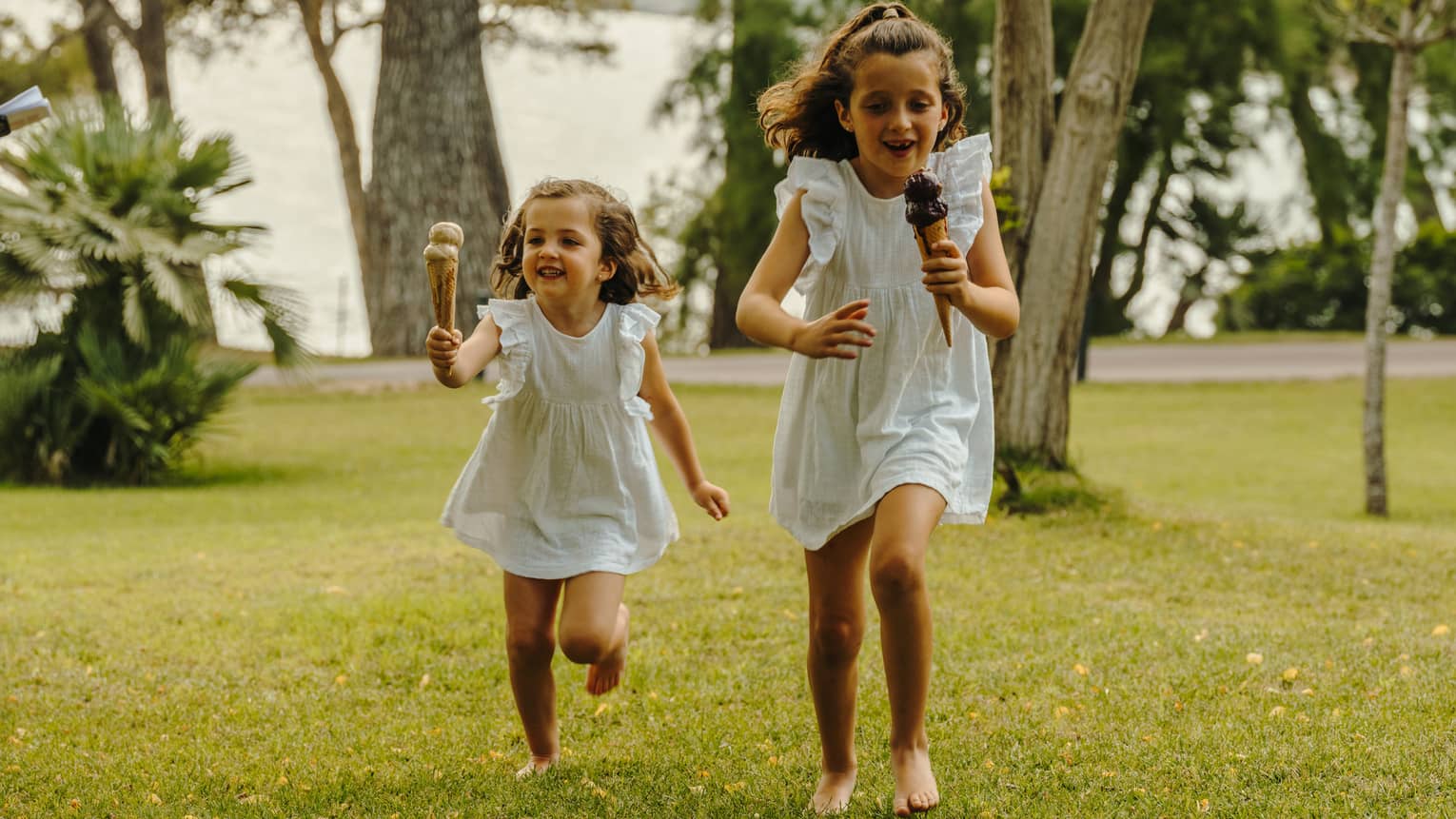 Two children holding double-scoop ice cream cones run across a verdant lawn dotted with trees.