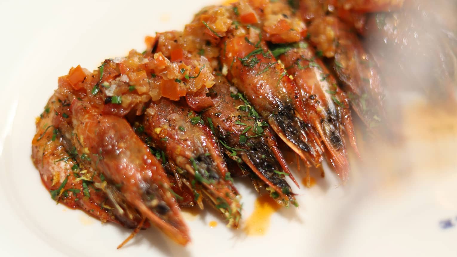 Cooked shrimp lined up in a row topped with tomato, herbs, onion and a garlic-lemon butter sauce