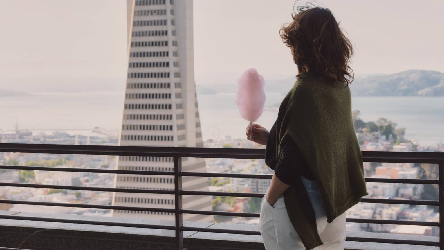 A person holding a stick of cotton candy stands on building balcony high above the city and looks out toward a skyscraper and a bay.