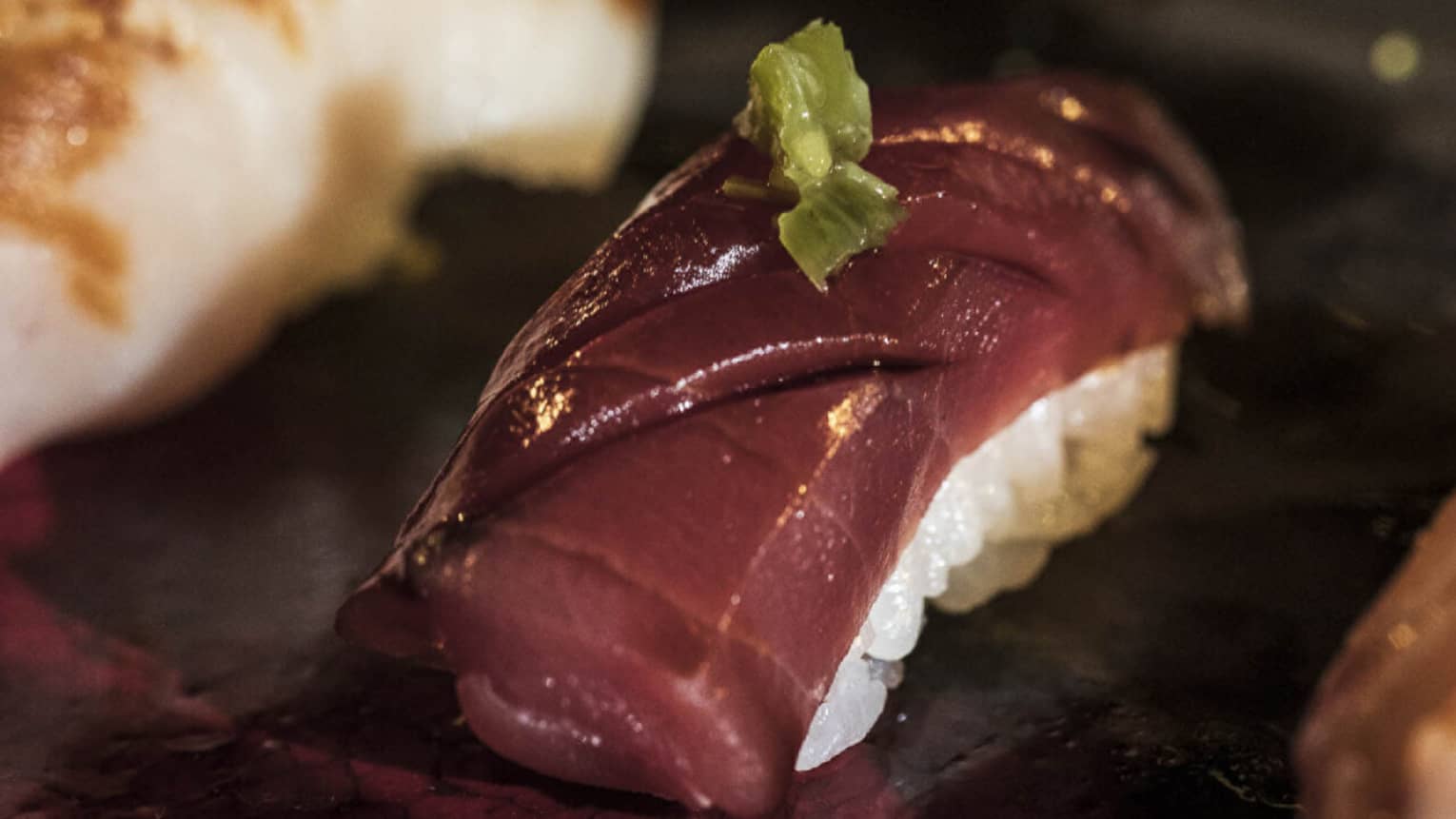Close up of a piece of nigiri on a plate. A white rice base is topped with a slice of deep-red fish and a small green garnish.