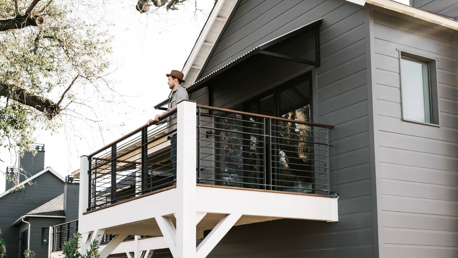 A man standing on a balcony of a grey building while looking at tree branches.