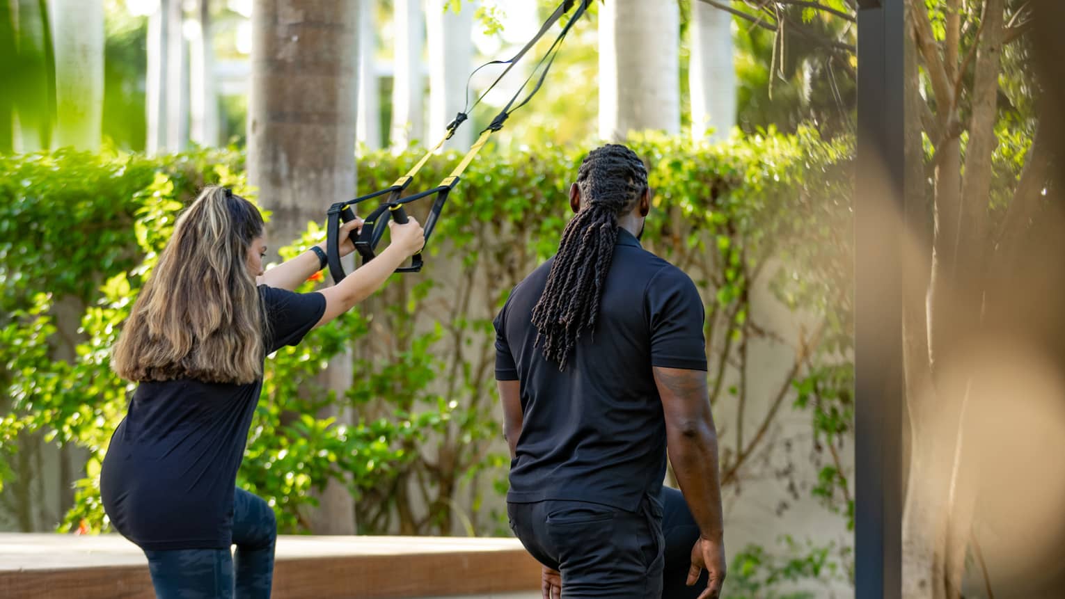 A guest practices lunging with suspended resistance bands in an outdoor, tree-shaded gym space. alongside a trainer.