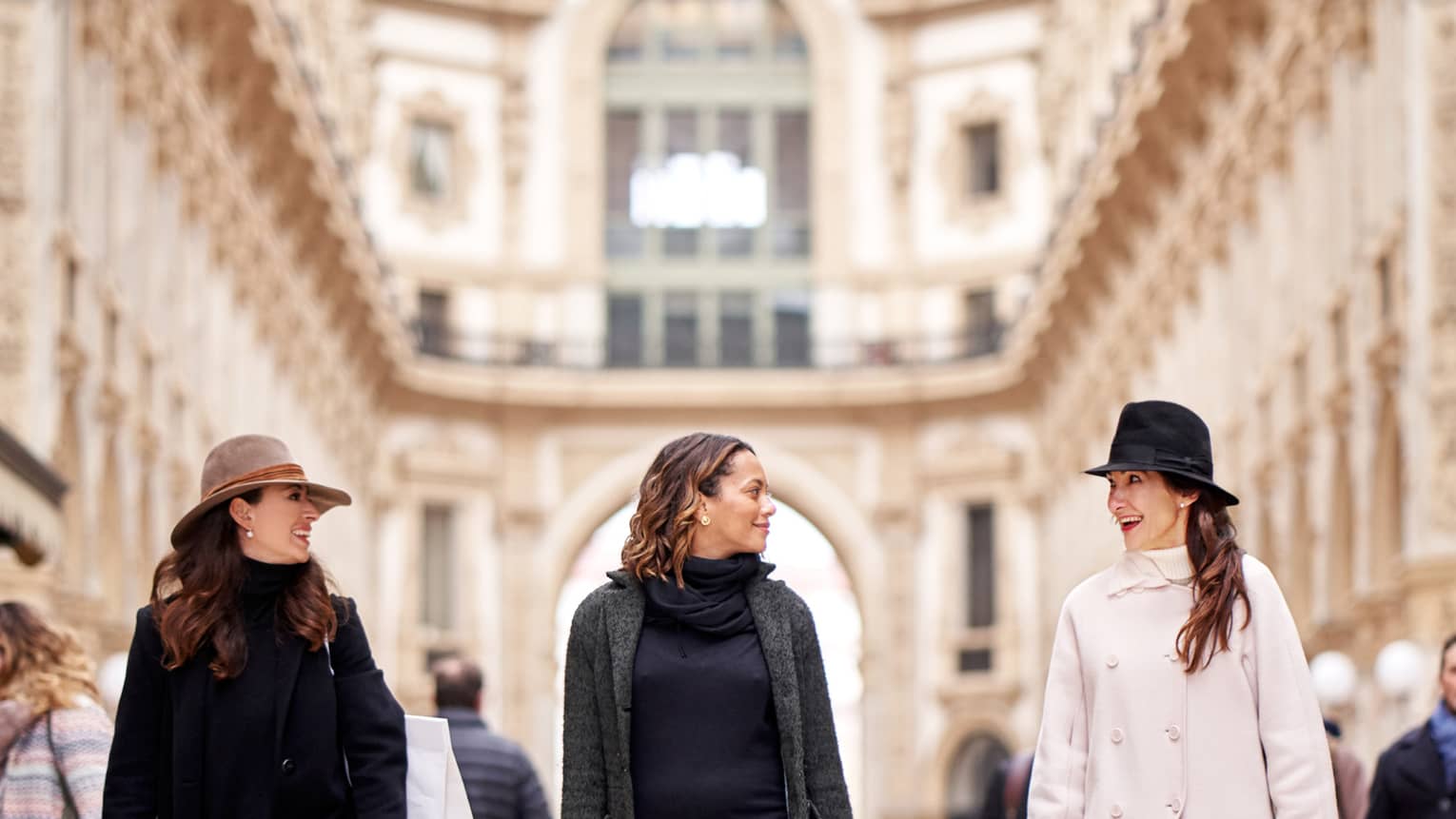 Front view of three chic shoppers smiling at each other as they stroll through the airy arcade of the shopping galleria.