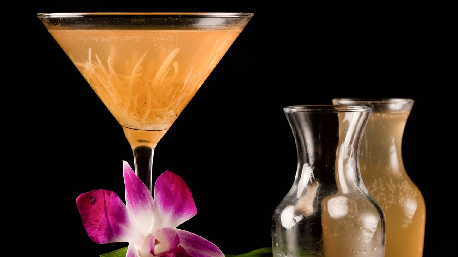 An orange martini glass on a wooden table surrounded by several pitchers of alcohol.