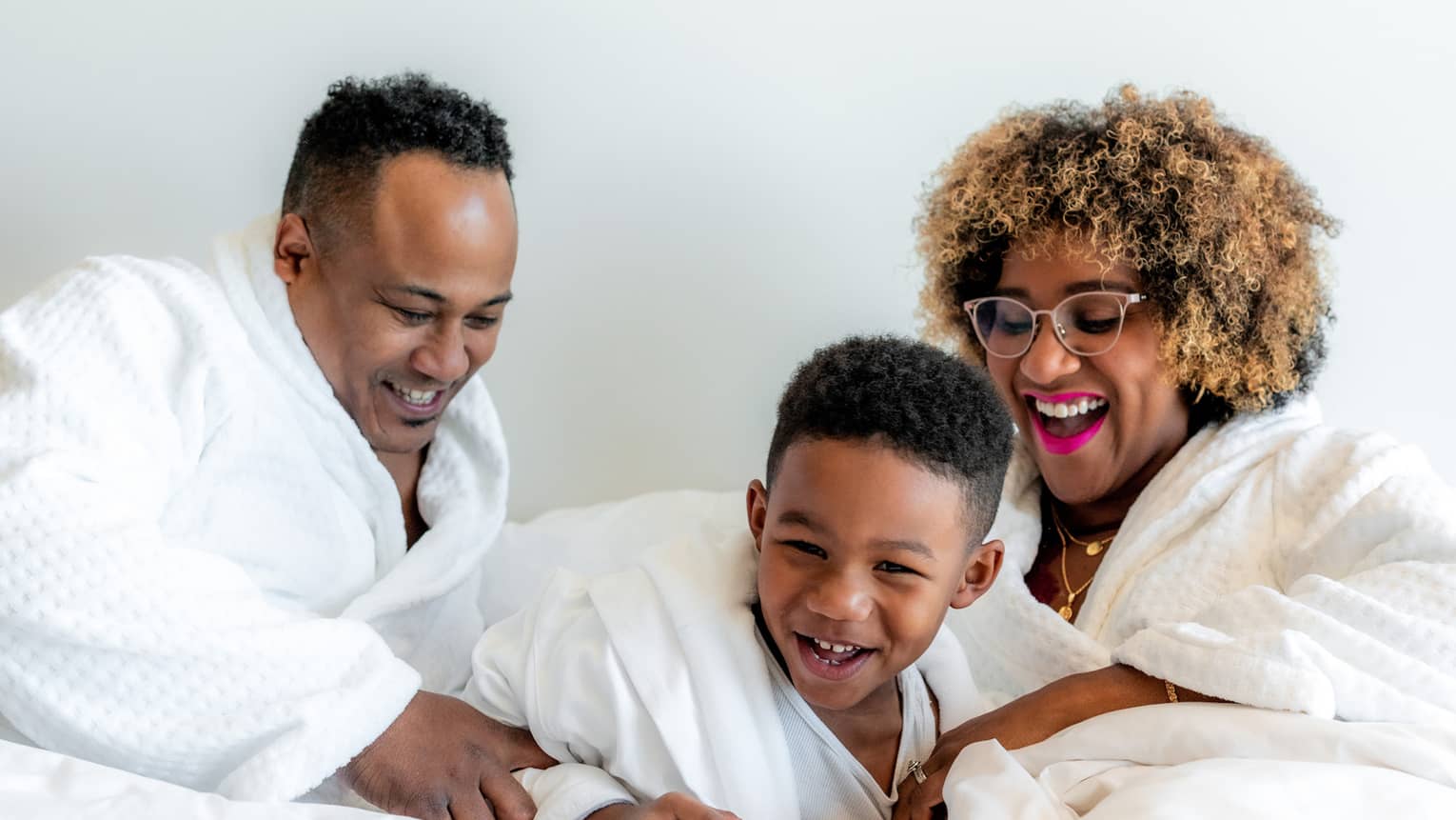 A mother, father and son playing on a white bed.