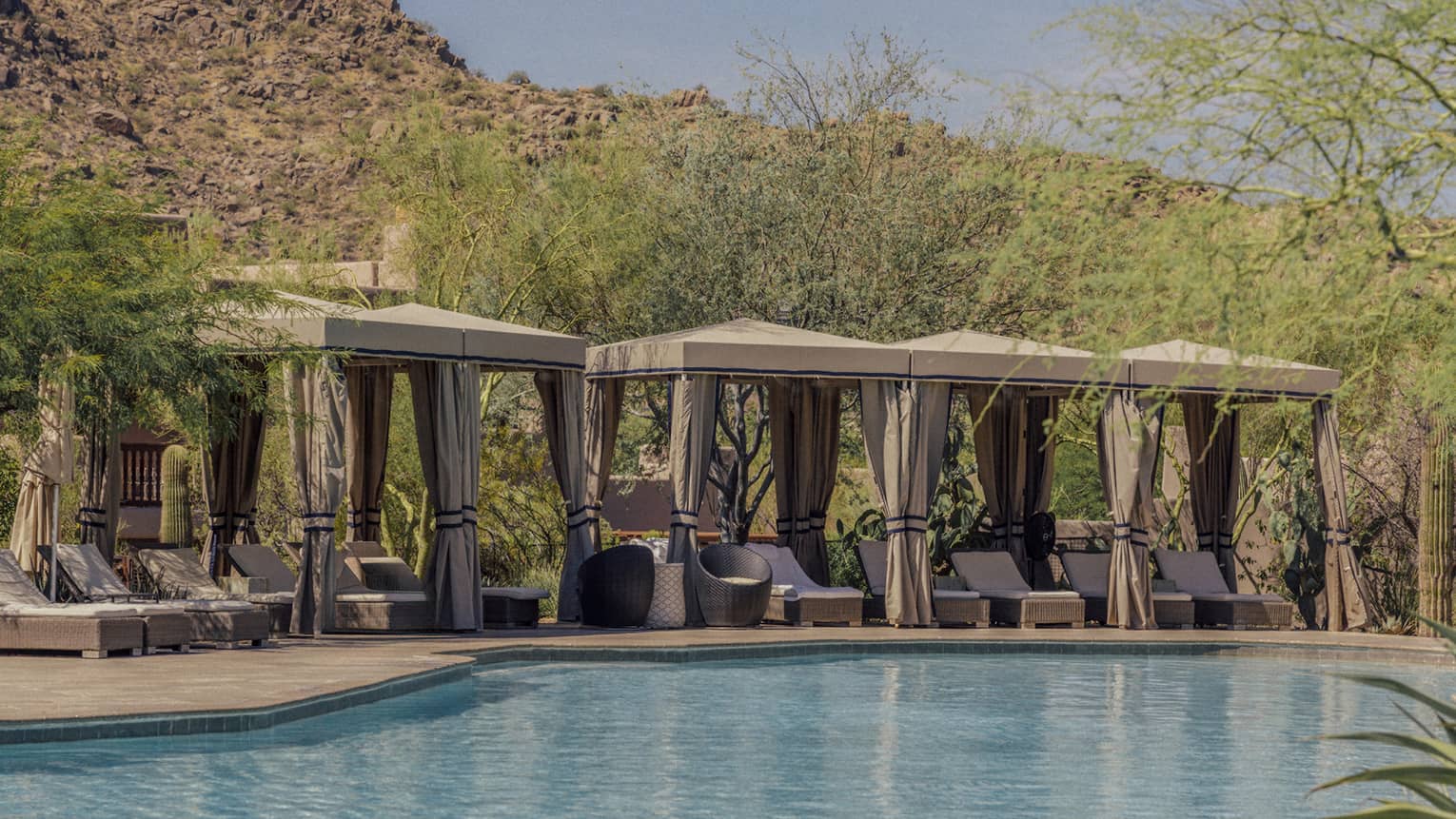 Four cabanas with lounge chairs near the pool with mountains in the background.