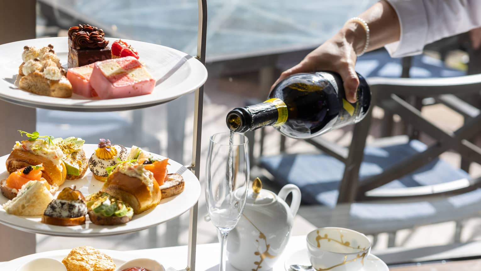 Champagne being poured next to a tiered assortment of afternoon tea small foods and pastries.
