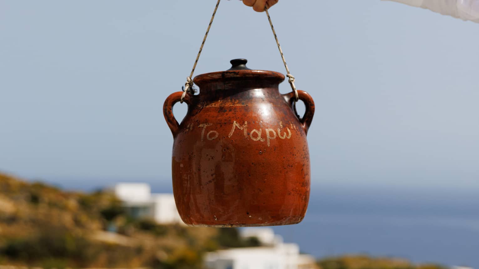 A glossy terracotta pot is dangled by a rope tied to its handles in front of an out-of-focus sky, sea and white houses.