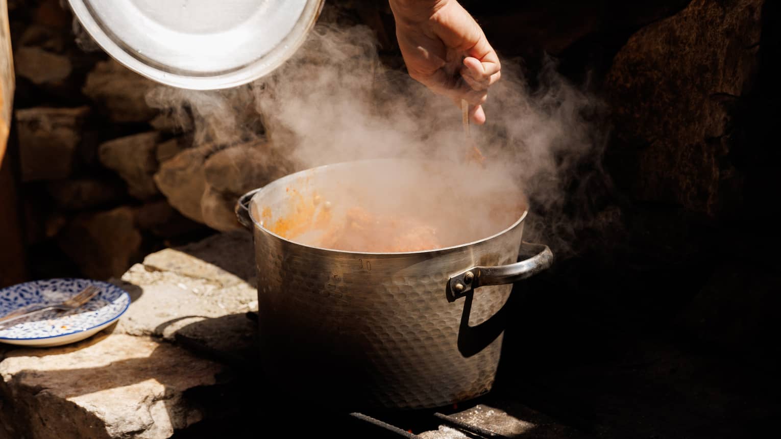 A dimpled metal pot on a stone cooking surface lets off a cloud of steam as a hand stirs the contents with a fork.