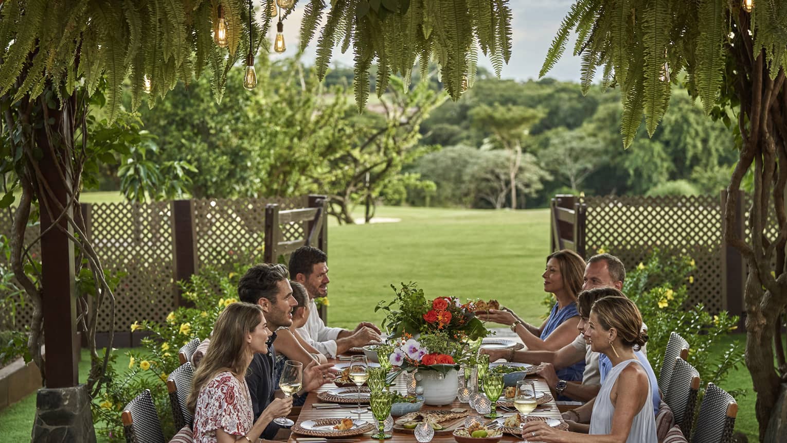 Eight people sit around a long dining table set up outside beneath a canopy of greenery and string lights