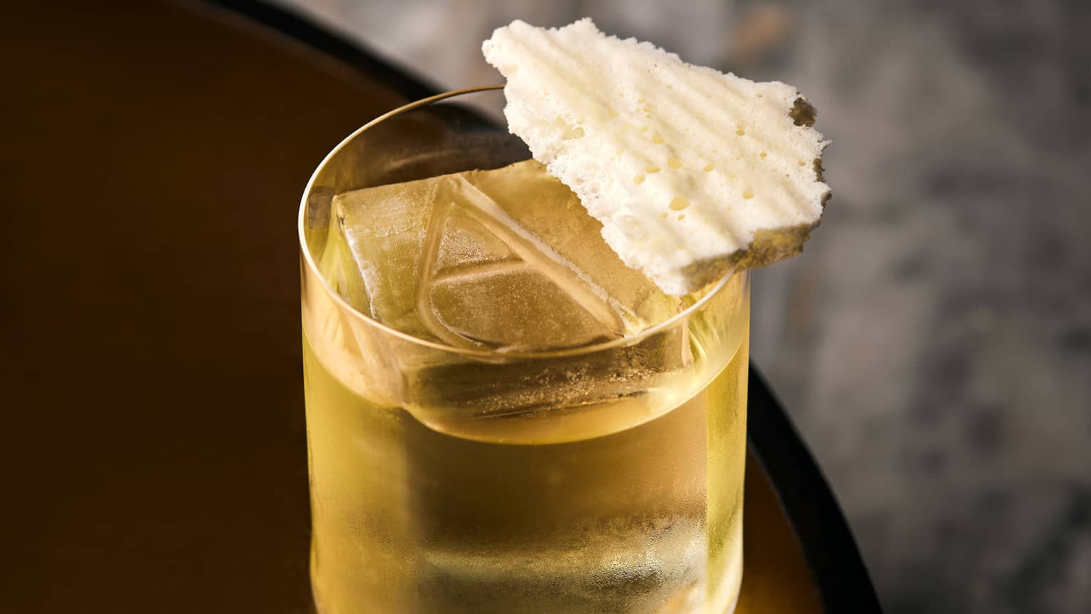 Yellow cocktail in a low glass with a large cube of ice, engraved with the letter A