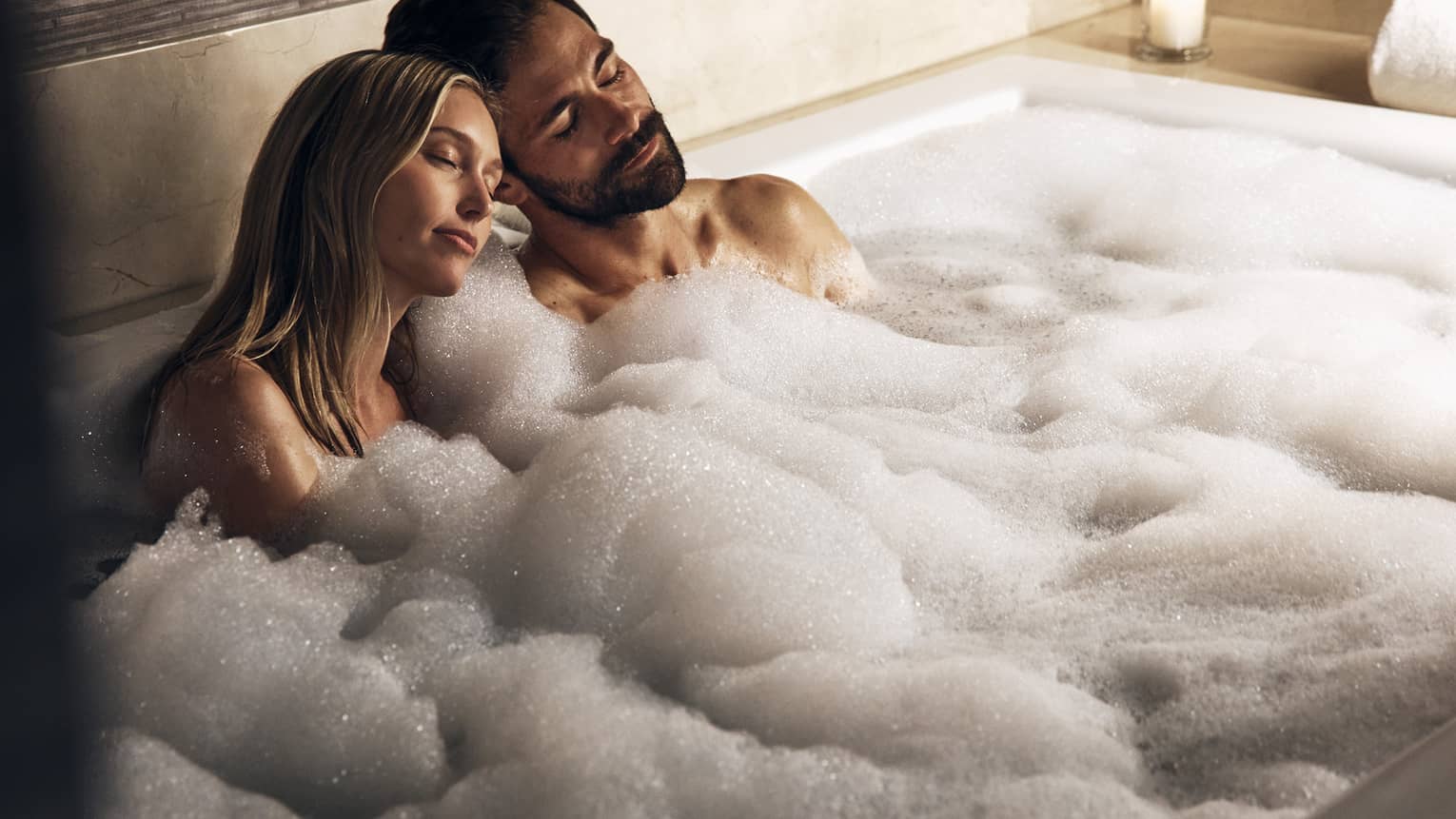 A couple sits together in a bubble bath with lit candles surrounding them, their heads leaning against eachother, both with their eyes closed