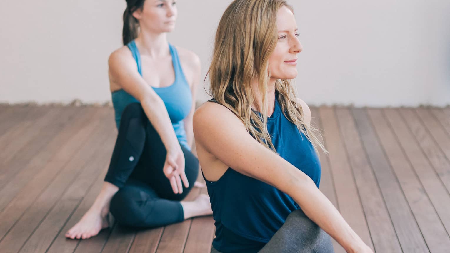 One blonde woman and one brunette woman sit with one leg crossed over the other with their bodies turned to one side and their front hands forming a lam mudra
