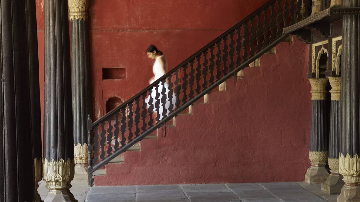 Side view of a guest descending a stone staircase, in a room supported by pillars with intricate gilded embellishments.