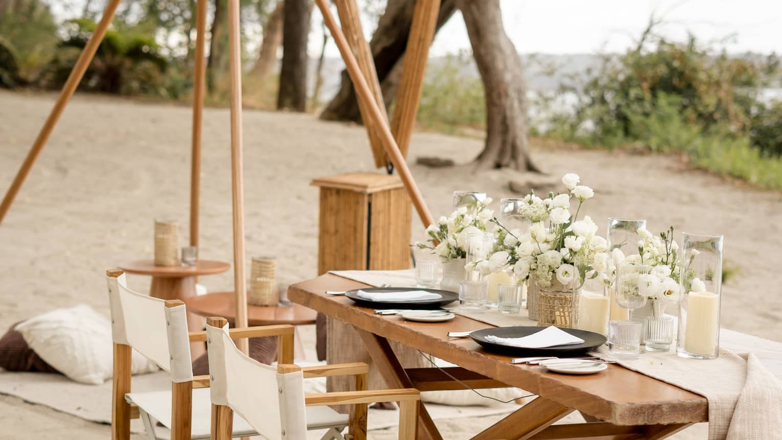 Wooden dining table and chairs set up on a beach, decorated with white floral centrepieces and candles