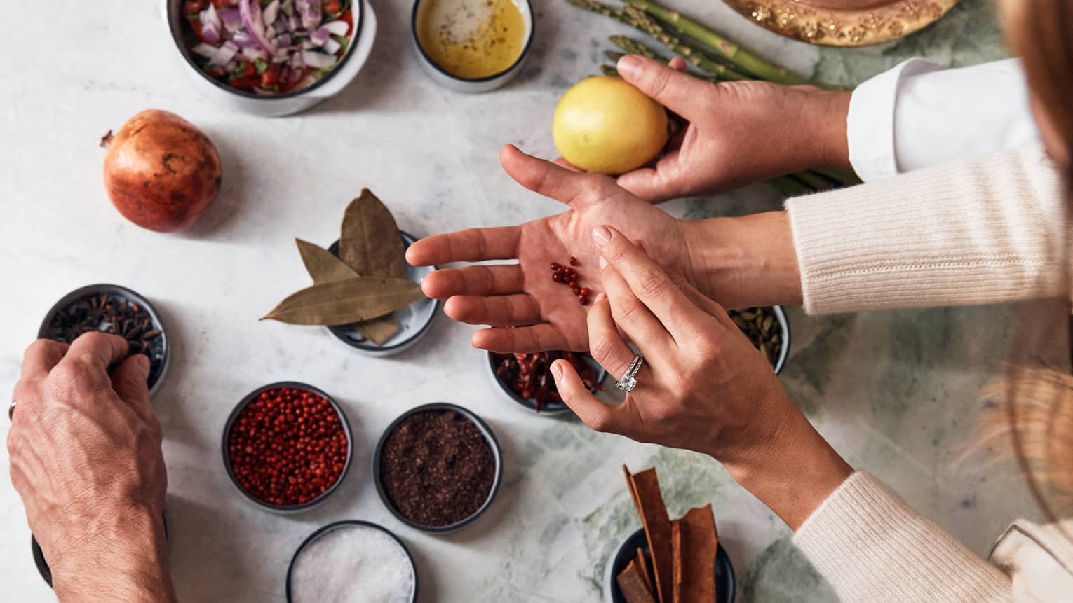 Thre pairs of hands reach over a marble table filled with various small bowls of spices and ingredients