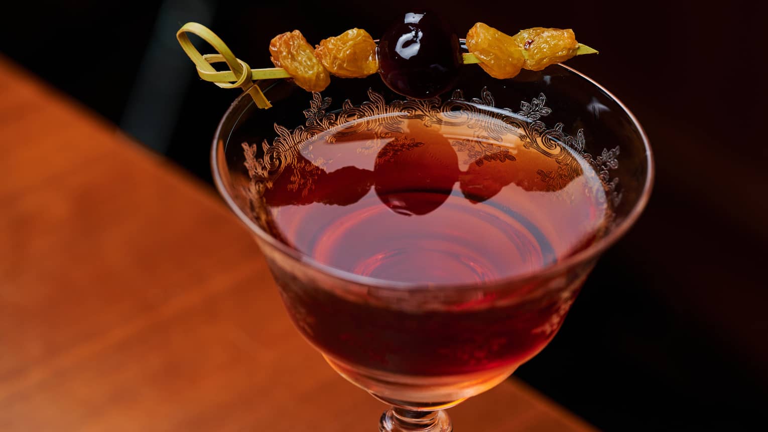 Close-up of a red cocktail in a finely engraved stemmed glass garnished with a skewer of raisins and a brandy cherry.