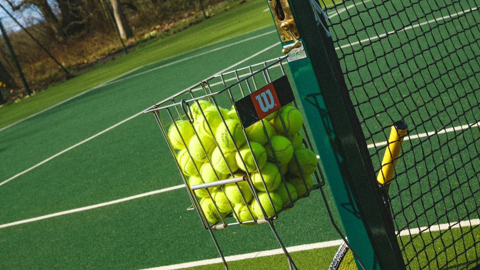 Tilted close-up of a basket filled with yellow tennis balls beside a net upon which leans a racquet on a green court.