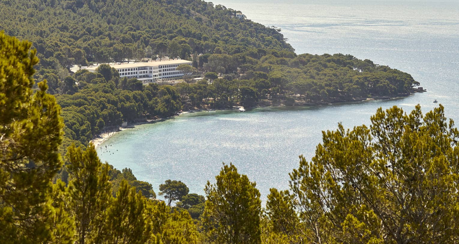 Hilltop view of a resort nestled among trees overlooking a shimmering bay dotted with sailboats and wide ocean vista beyond.