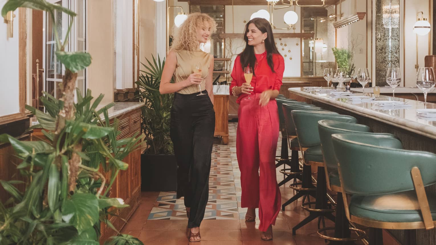 Two women walking a well lit restaurant with colourful tiles and seat at a bar.