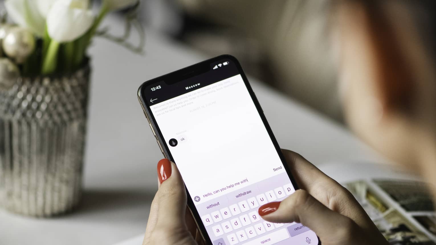 Woman holds a smartphone, typing. A book and vase of flowers lie on the table.