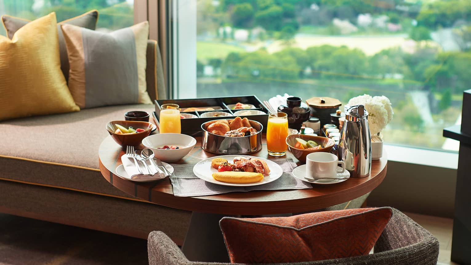 A breakfast tray of pastries, fresh fruit, juice and coffee on a guest room lounge chair at a window overlooking lush gardens.