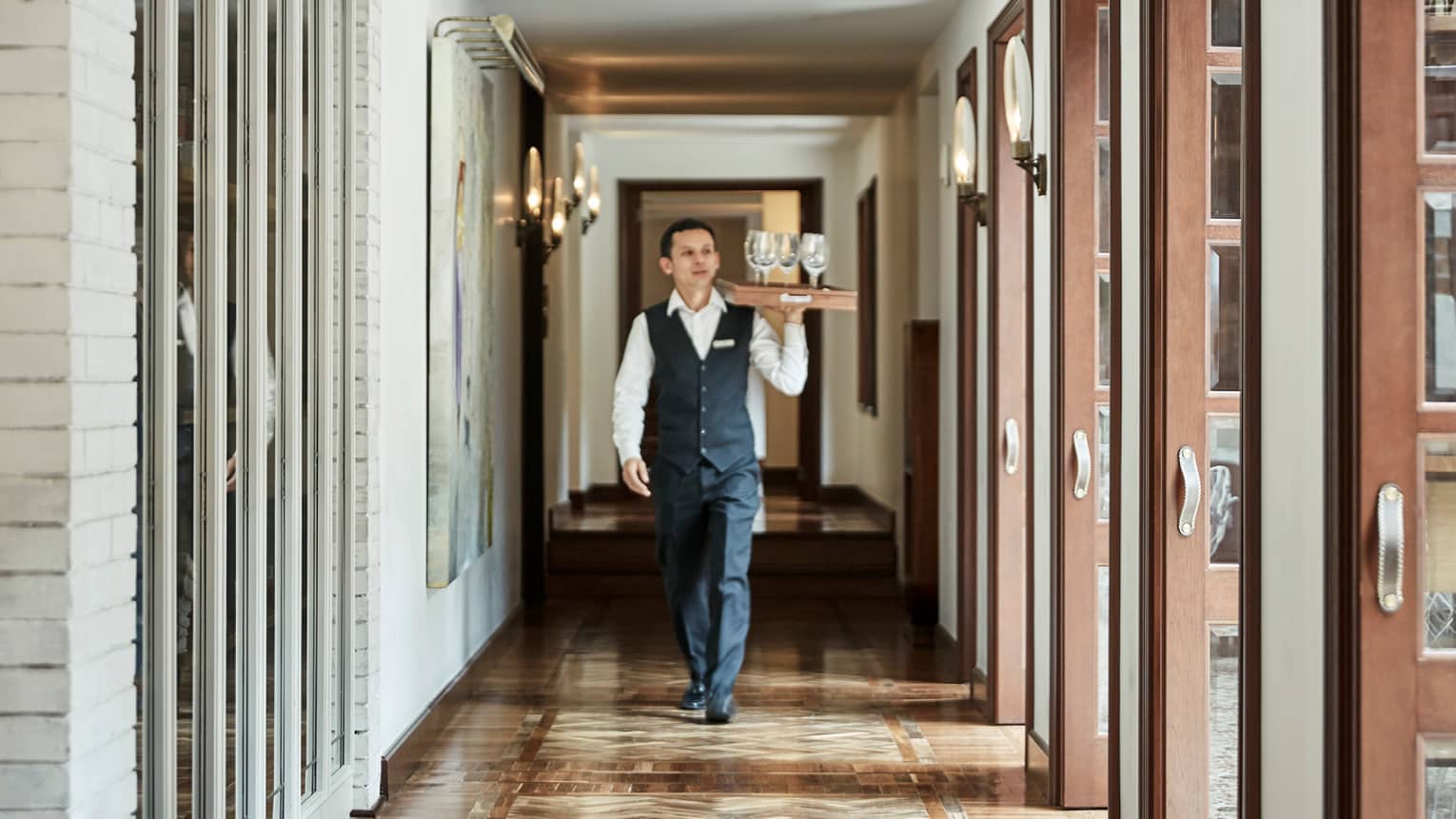 Server in black vest and pants, white shirt, carries tray with wine glasses down sunny hall with wood floors