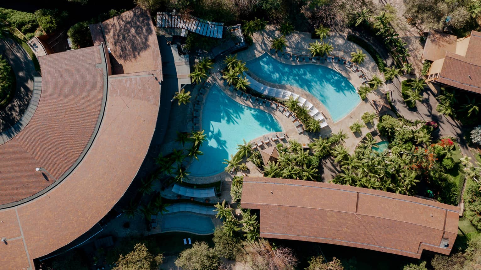 Aerial view of two pools nestled between buildings in a narrow strip of forest with sand on either side