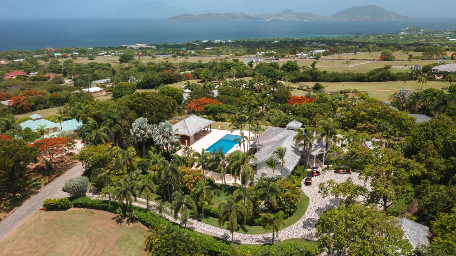 Aerial view of private villa with pool, palm-tree lined driveway, ocean in distance