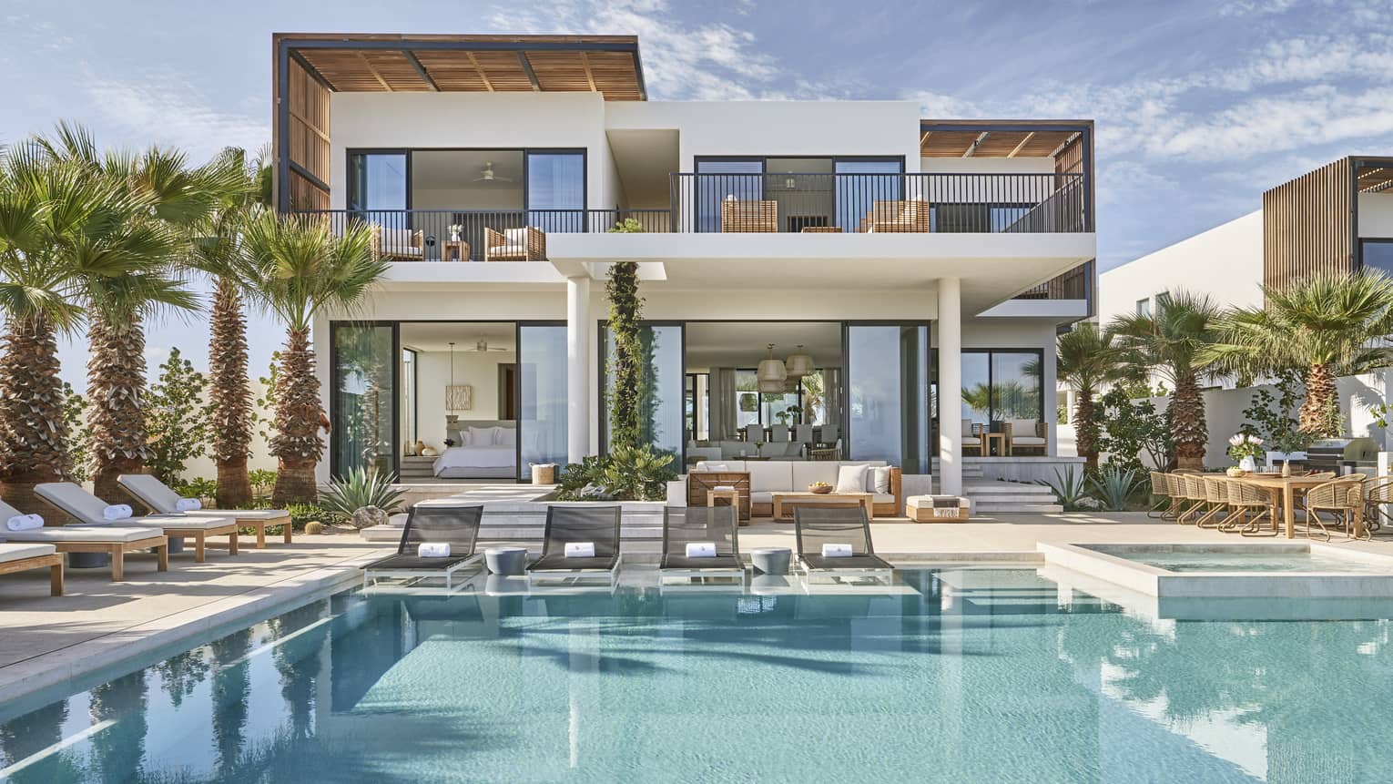 Modern luxury villa with a spacious pool area, surrounded by palm trees, featuring outdoor lounge chairs, a dining area, and floor-to-ceiling windows.