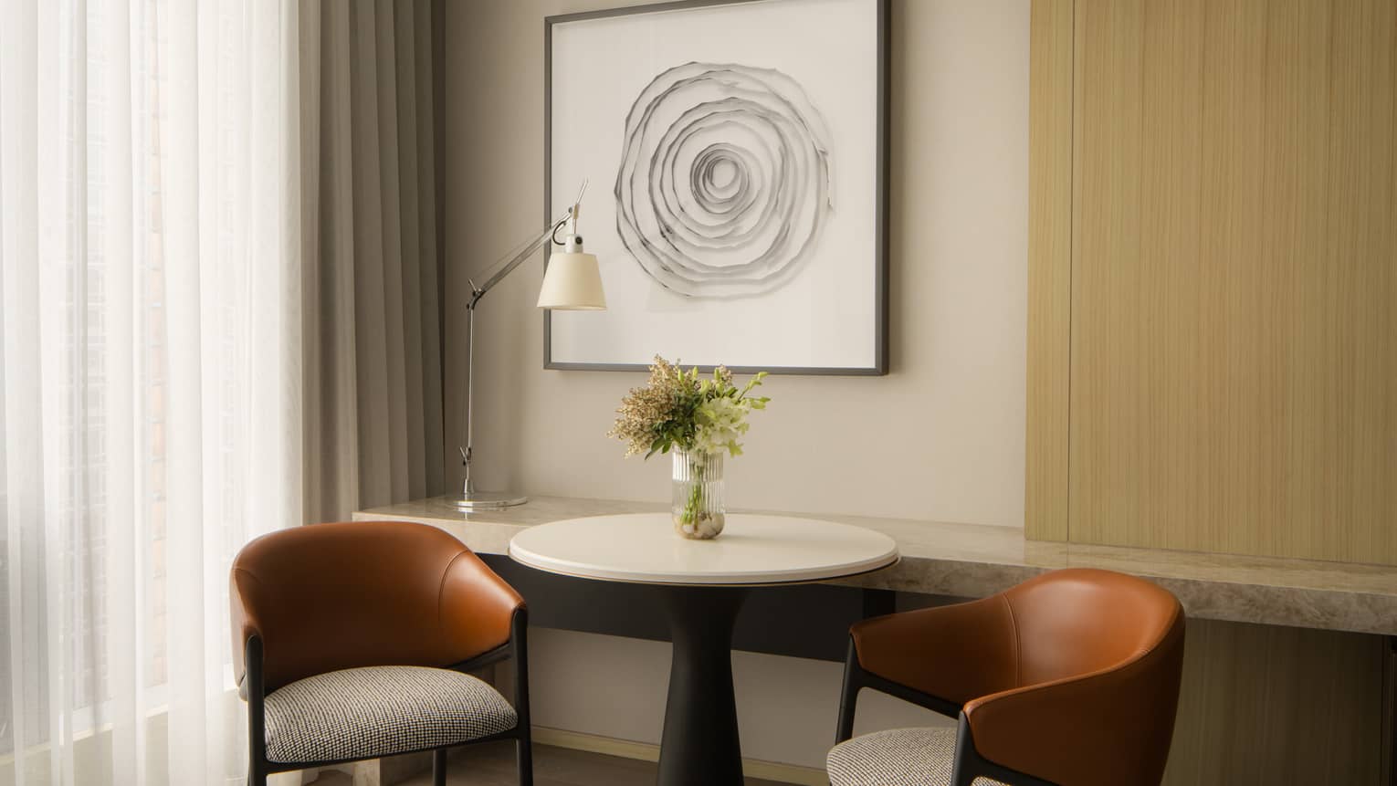 Cozy seating area with two leather chairs, a round table with a vase of flowers, and abstract wall art in a hotel room