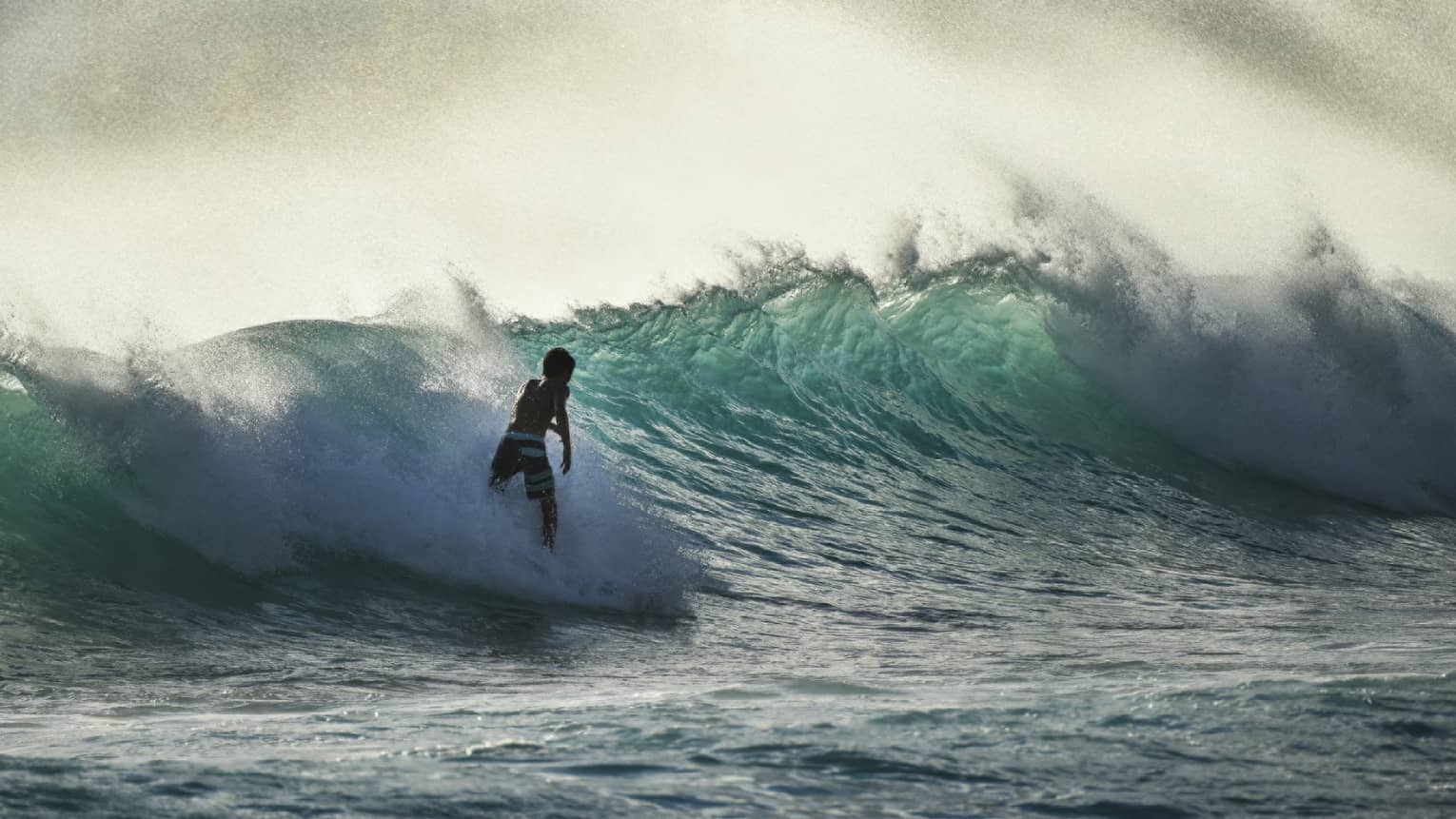A surfer silhouetted in a spray of sea-foam rides a large wave in a teal-coloured ocean under a misty, yellow-streaked sky.