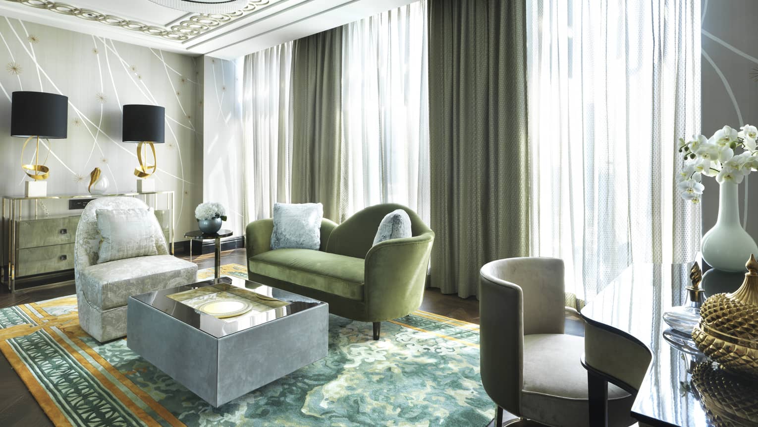 Luxurious seating area with green velvet sofa, elegant armchairs, mirrored coffee table and gold-accented lamps, framed by soft drapes and natural light
