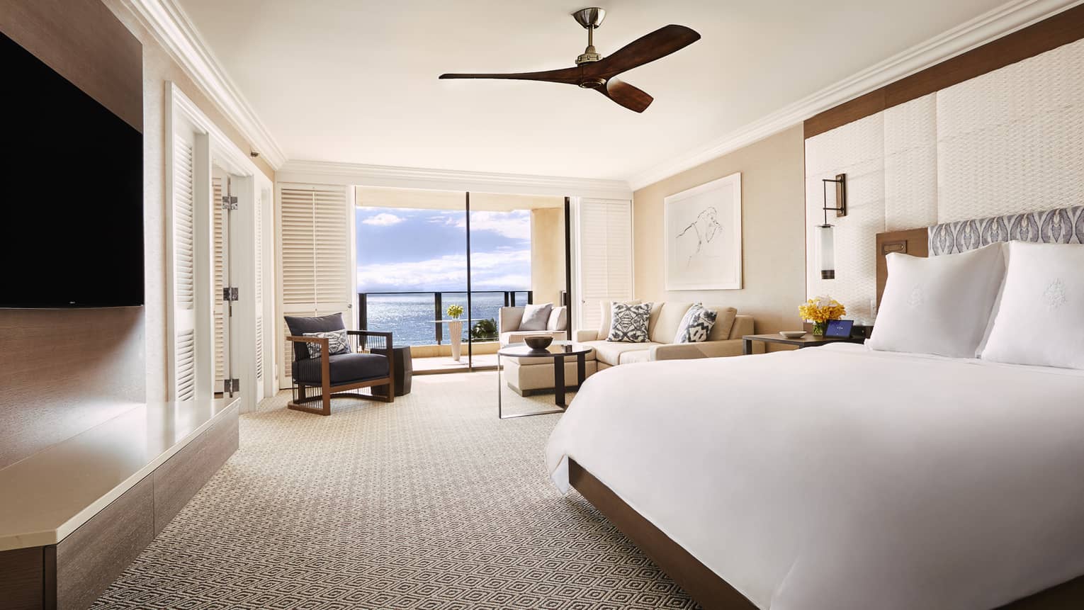 Bright suite with king-size bed, seating area and ocean-view balcony, featuring neutral tones and a ceiling fan at a Four Seasons luxury resort