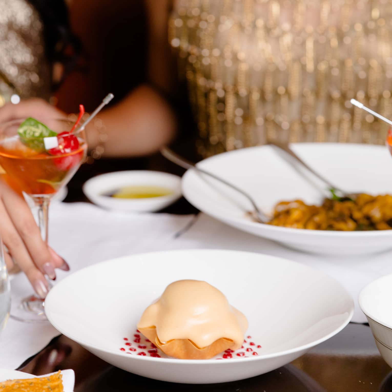 A woman in a gold sequin dress sits at dining table with dishes in front of her and red cocktail