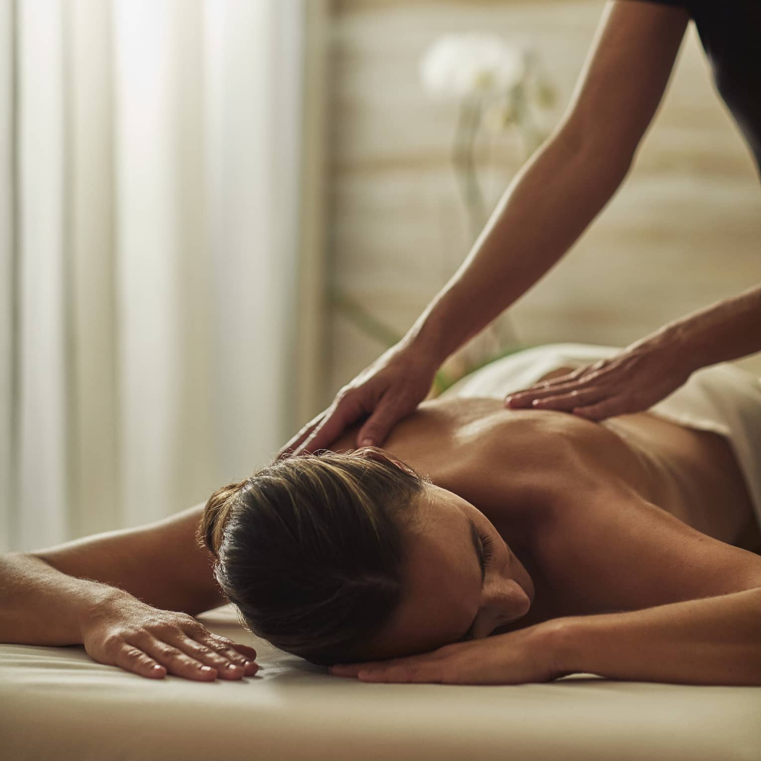 Woman in black uniform massages bare shoulders of woman lying on table face-down with head to side