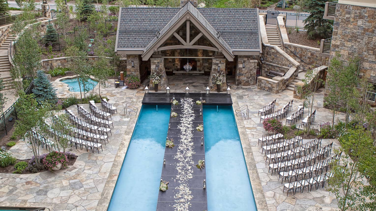 Outdoor wedding setup with a long black aisle over a pool, lined with flower petals, leading to a stone chapel. Surrounded by trees and mountains. 