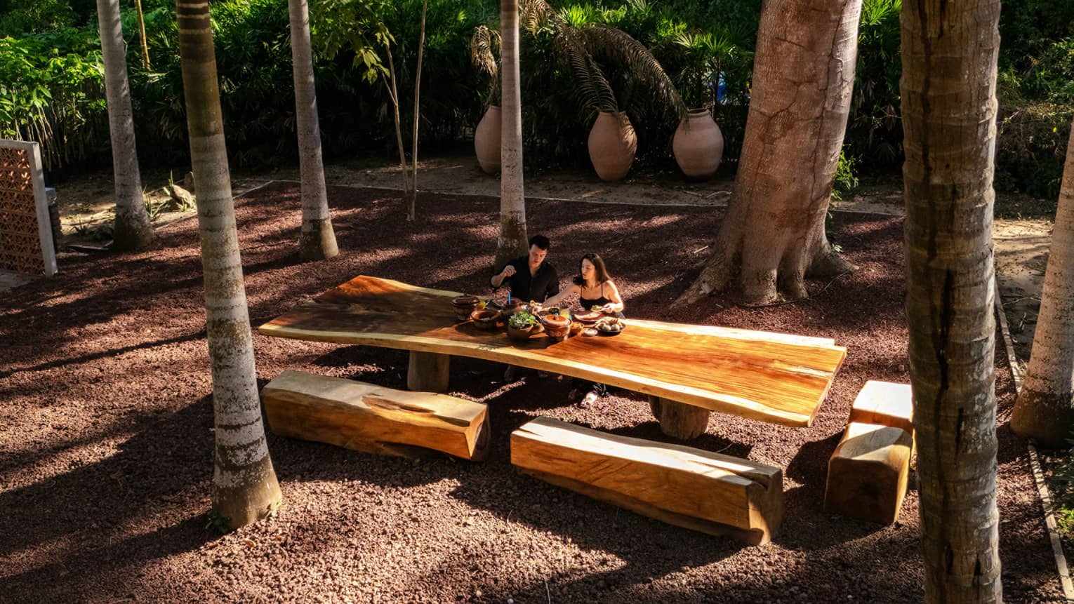 Two people sit at a long wood outdoor table with wood beam bench seats, all surrounded by palm trees.