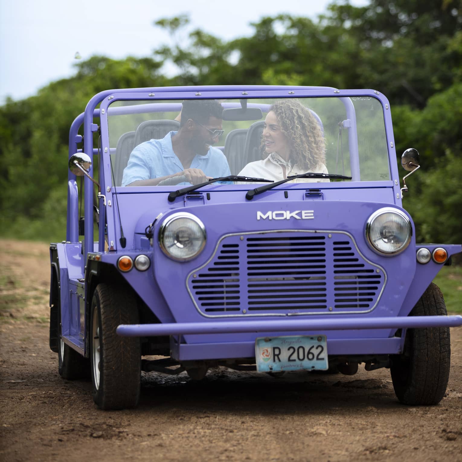 Front view of a couple driving a purple Moke, a small open-air recreational vehicle, along a dirt road surrounded by trees.