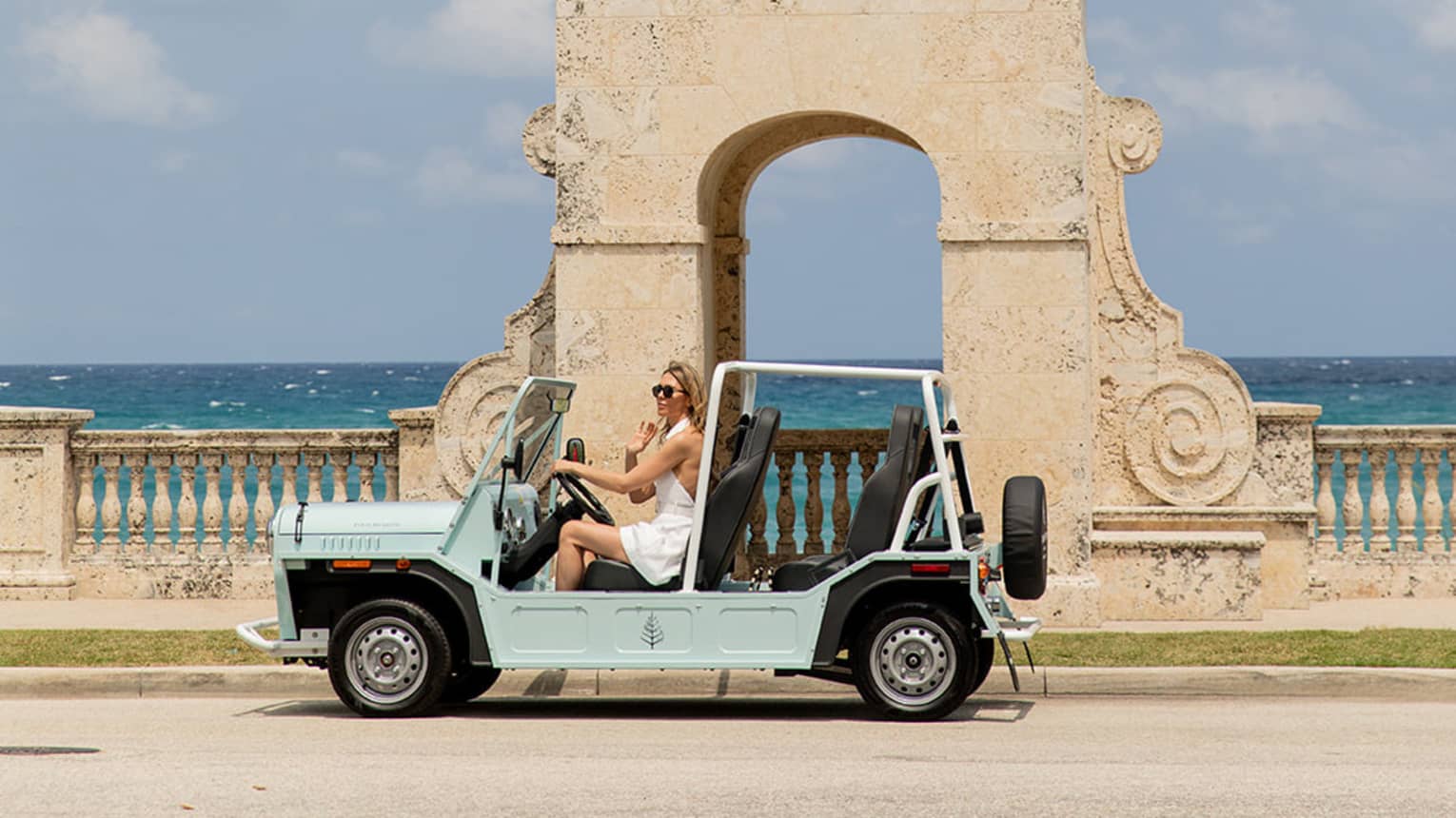 A person drives a light blue open-top vehicle past a historic clock tower by the sea. The backdrop features clear skies and the ocean.