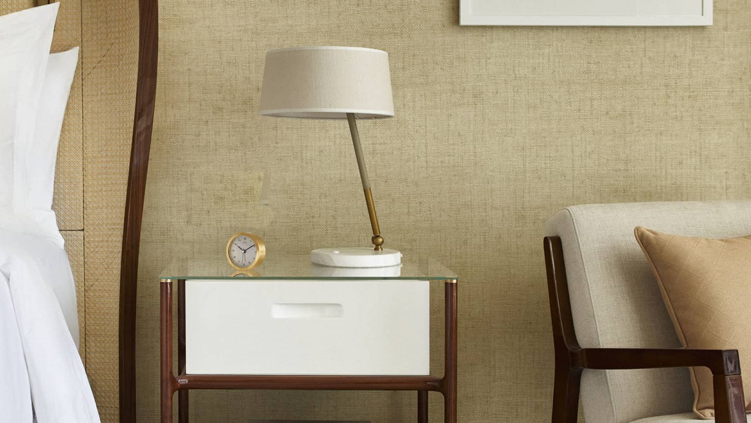 Next to a bed sits a nightstand with lamp, arm chair, square framed artwork on wall