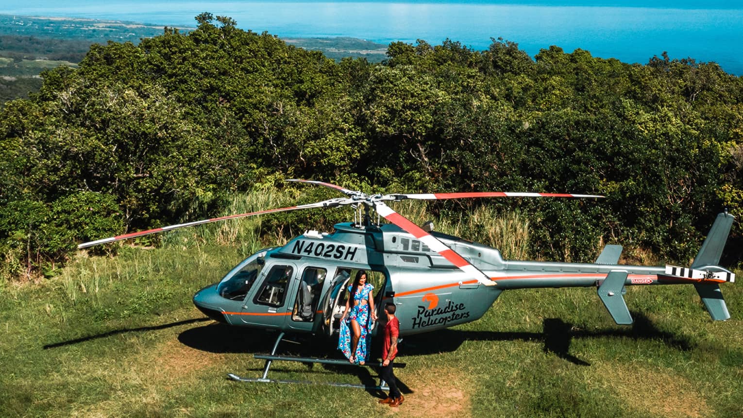 Woman and boy standing beside helicopter on grass with bushes and ocean water behind them
