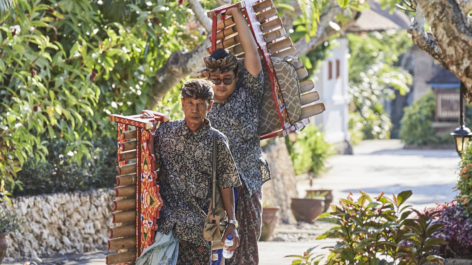 Two musicians in sarongs and printed shirts carry colourful bamboo xylophones down a stone path lined with lush foliage.