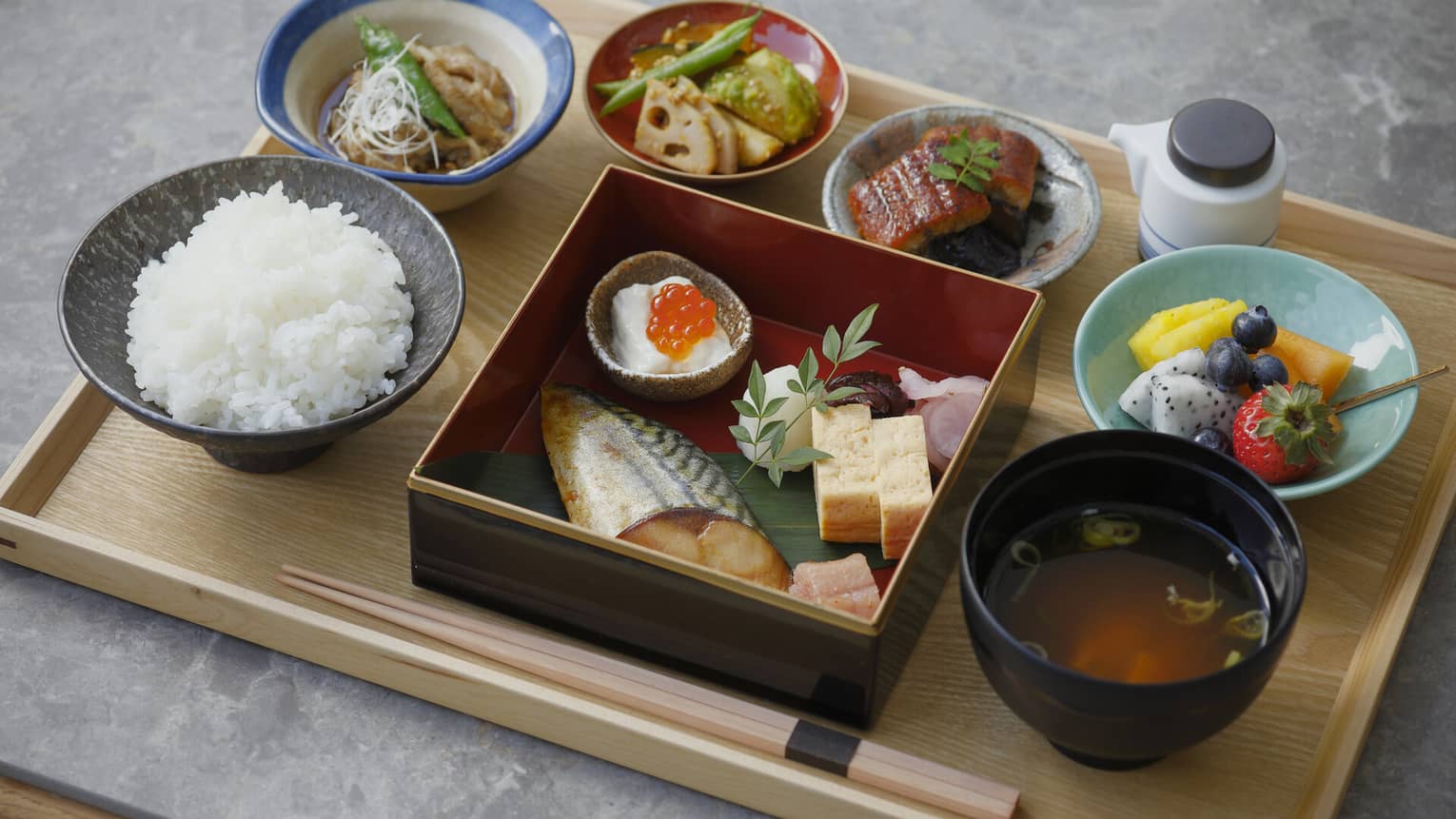 A close up of a Japanese breakfast on a natural wooden tray ? fish, chopsticks, rice, soup and fruit