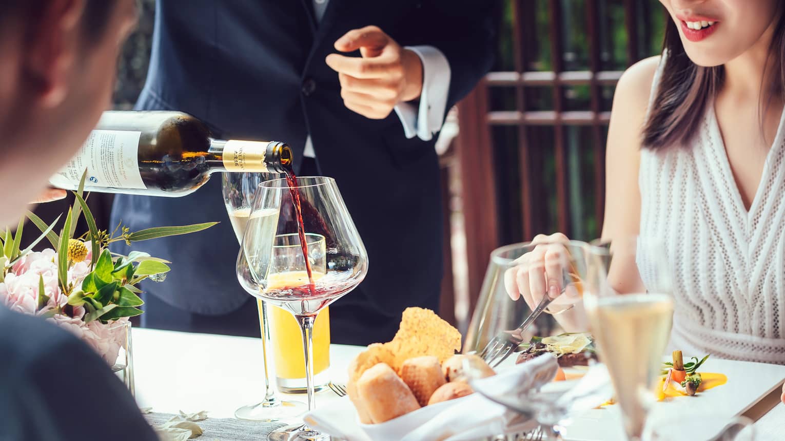 Server pouring red wine into large glass as couple dines at table