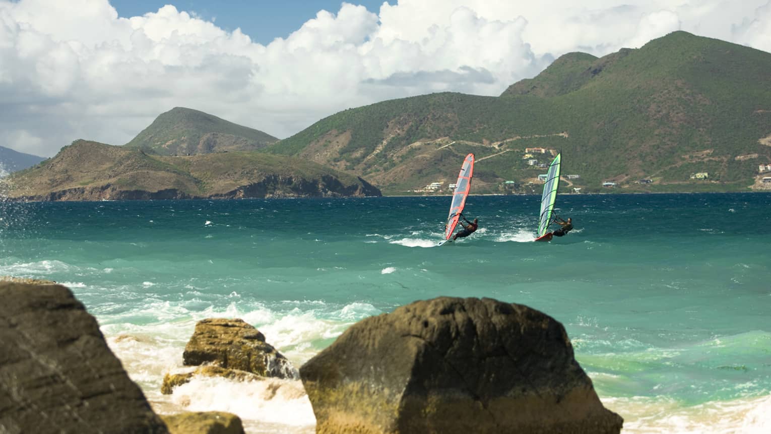 Two windsurfers glide on waves in bay between rocks, green mountains