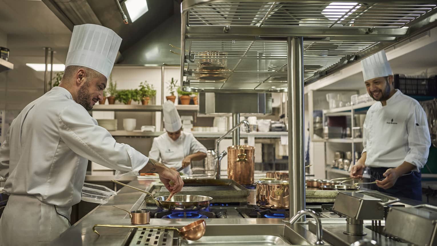 Three chefs with white coats and chefs hats cook in a kitchen