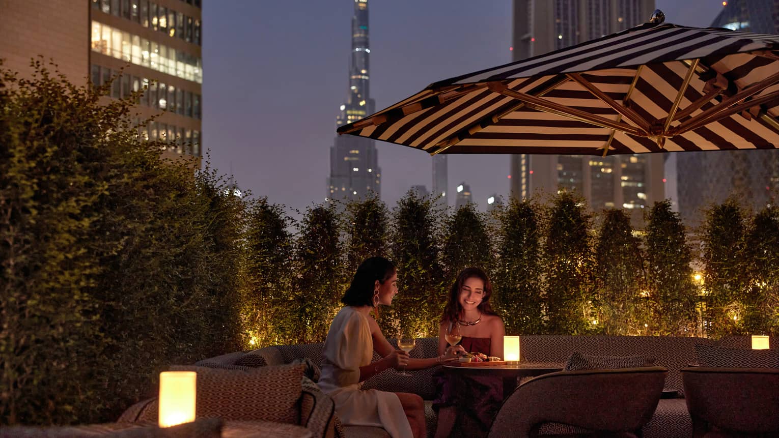 Two women sit at an outdoor table at dusk with a candle in the middle and a patio umbrella overhead