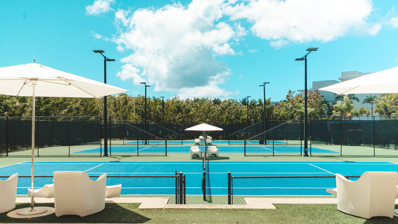 Three turquoise tennis courts with low armchairs and shady umbrellas on the sidelines and lush trees around the perimeter.